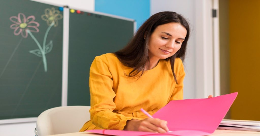 A woman seated at a desk with a pink folder, preparing nursery report card comments and behavior remarks for children.