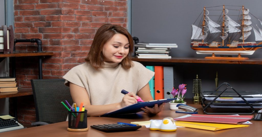 A teacher at a desk writing nursery report card comments for behavior while holding a clipboard.